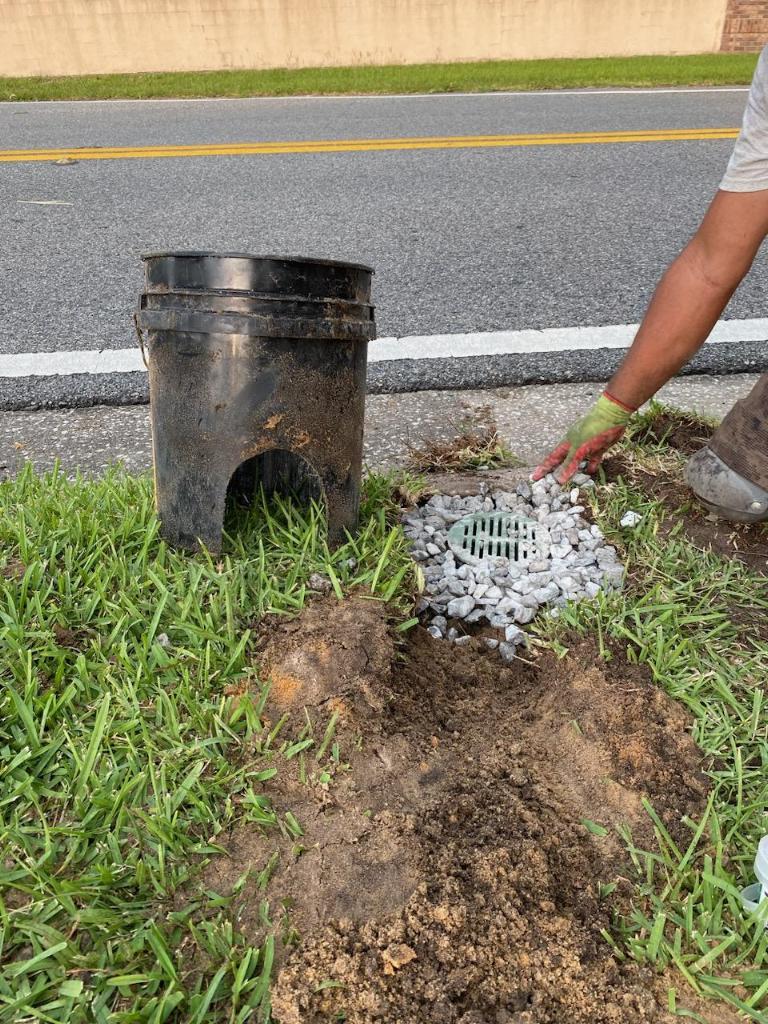 Worker installing a gravel-covered drain grate beside a black bucket catch basin near a curb